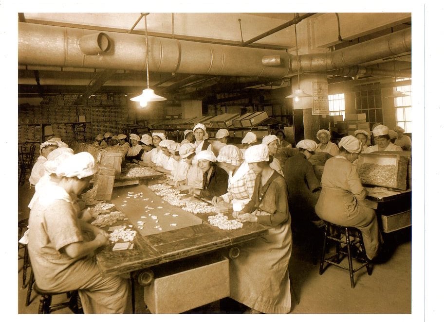 Female employees wrapping candies at large tables in a factory for Wallace Candies in 1914.
