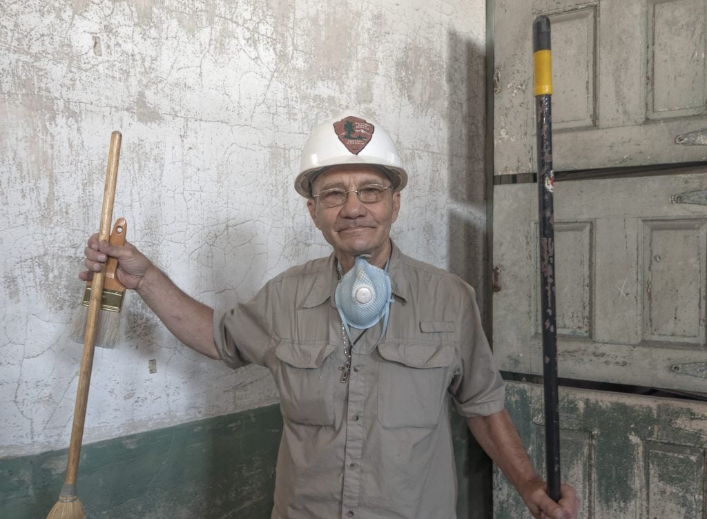 A man in a hard hat smiles, holding a crooked broom