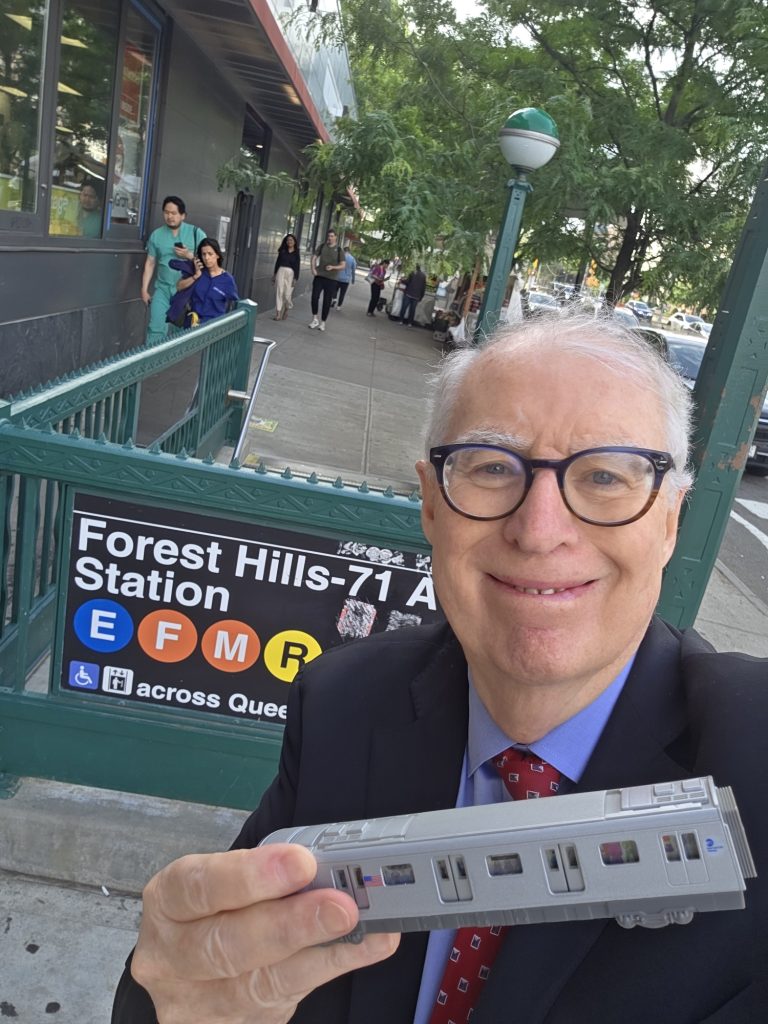 Charlie Pellett in front of the Forest Hills-71St Street Station holding a toy subway car