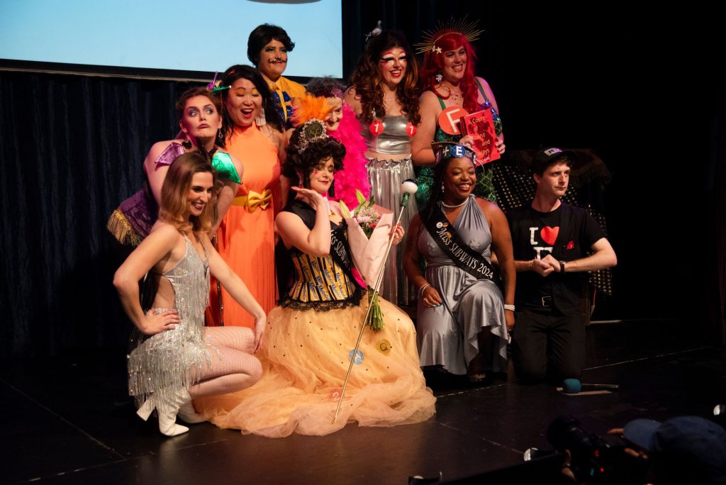Miss Subways contestants, in colorful and fancy dress, pose on stage.