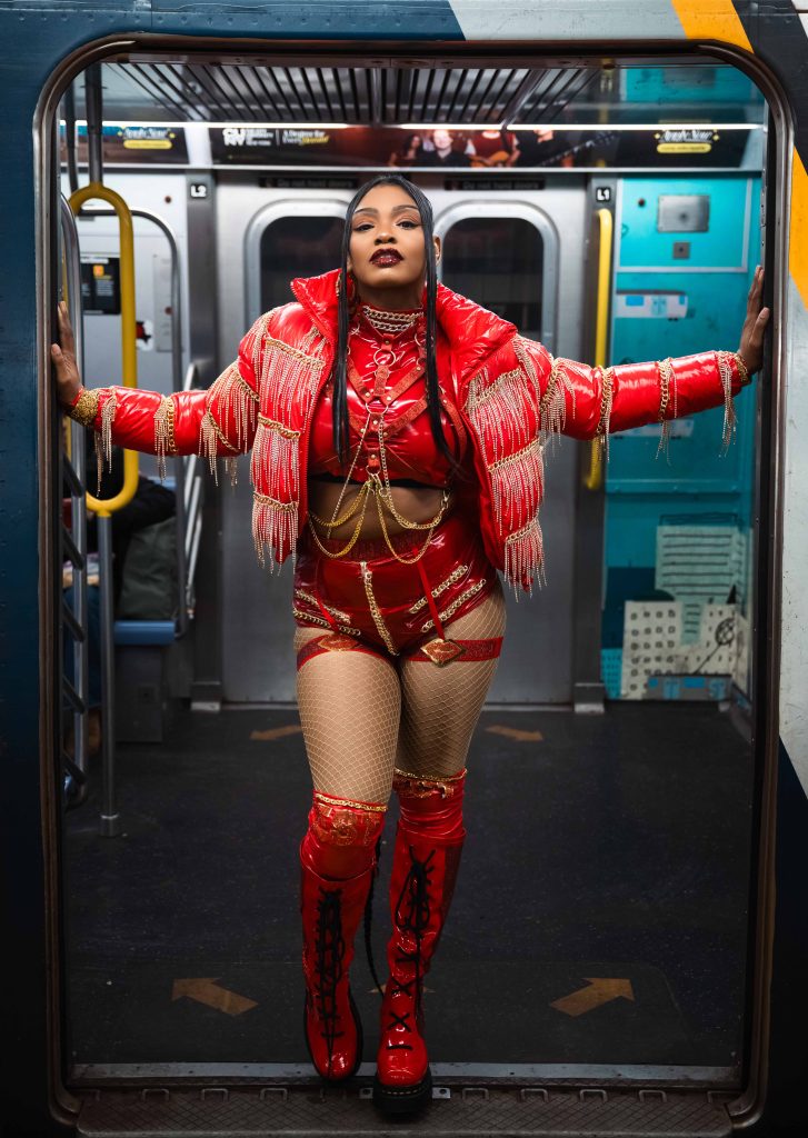 LuXury Leah on a subway train in a red outfit with chains and fringe.