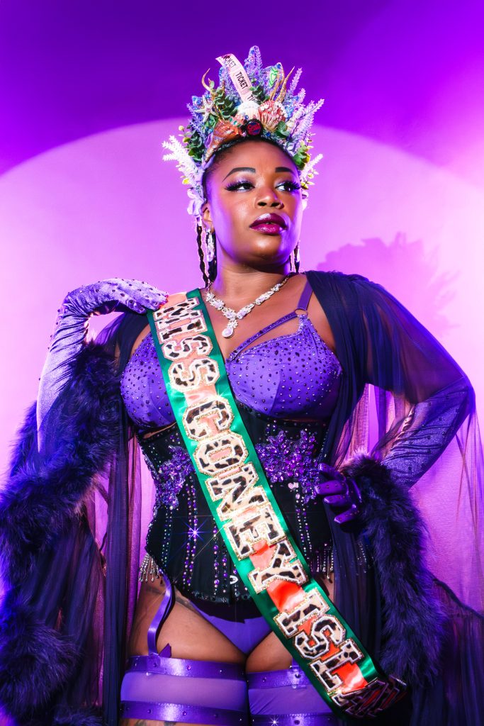 Queerly Femmetastic in a purple negligee and Miss Coney Island crown and sash